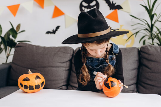 Close up of cute little girl preparing for Halloween, painting drawing face on pumpkin while sit on comfy sofa in child room at home, adorable kid making jack-o-lantern. Holiday decoration concept