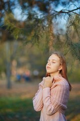 Portrait of a girl in a knitted sweater in an autumn park among coniferous branches. September or October, yellow leaves on trees.