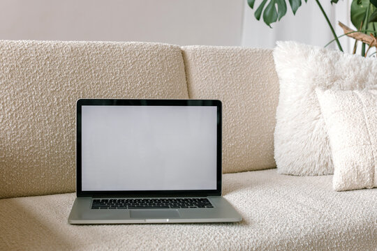 Laptop computer with blank screen on white sofa with pillows. Aesthetic template with mockup copy space. Online shopping, blog, social media, website branding, showcase. 