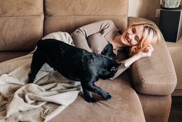 Happy woman play with their dog black french bulldog. Girl petting a puppy indoors in the stylish living room with plaid on the couch