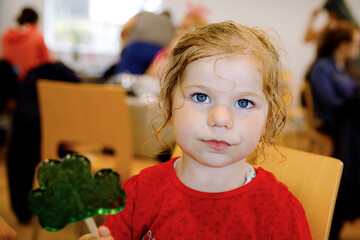 Cute toddler girl with cloverleaf lollipop in Irish pub or cafe. Child resting with parents and...