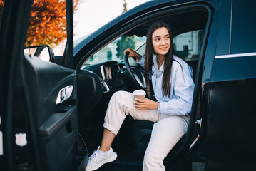 A happy attractive woman or businesswoman holding a cup of coffee and getting out of her modern car