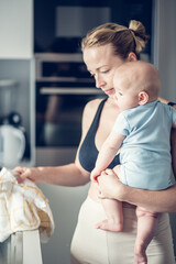 Woman wiping kitchen sink with a cloth after finishing washing the dishes while holding four months old baby boy in her hands