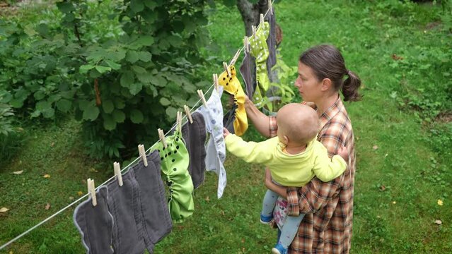 Top view mother with little baby together removes reusable diapers drying on clothesline in garden. Modern eco friendly cloth nappy for infant child hygiene. Zero waste concept.