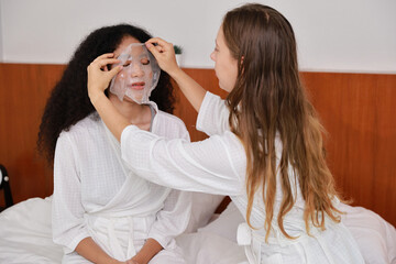 Young happy diversity ethnic beautiful woman in bathrobe sit on bed and doing cosmetic white facial mask for skin care and treatment. Women lesbian couple daily LGBTQ lifestyle and love concept.