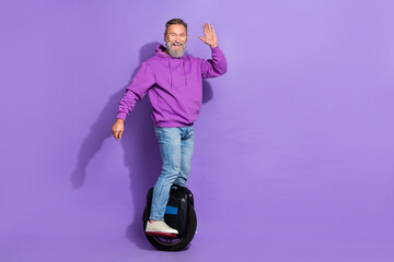 Photo of nice retired senior funky man dressed purple hoodie driving monowheel waving palm greetings isolated on violet color background © deagreez