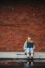 Fototapeta na wymiar Happy young curly woman student typing on laptop sitting outdoors in city street against brick wall background with copy space. Internet technology. Modern