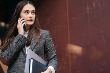 Confident business woman walking, holding textbook, laptop, talking on mobile phone on city street in front of modern office building. Copy space. Modern