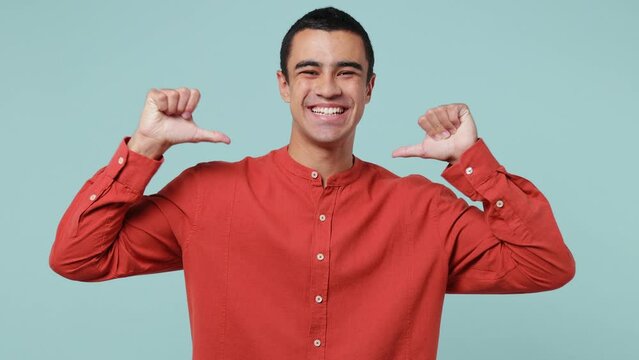 Young confident middle eastern man he wear red shirt pointing fingers on himself blinking showing thumb up like gesture isolated on plain pastel light blue cyan background. People lifestyle concept