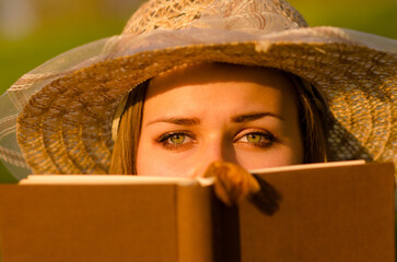 Young woman wearing dress and hat reading book in the nature on a sunny summer day