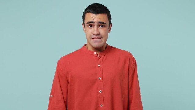 Young confused shy shamed middle eastern man 20s he wear red shirt fun look camera spread hands say i do not know oops ouch oh omg isolated on plain pastel light blue cyan background studio portrait