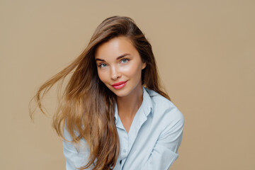 Fototapeta premium Portrait of attractive young woman has fair hair floating in wind, natural beauty, wears makeup, dressed in stylish shirt, isolated over brown background, has healthy skin. People, style, feminity
