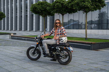 Shot of trendy guy with long hairs driving motorcycle outdoors in daytime.
