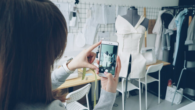 Young female clothing design blogger is shooting tailoring dummy with half-finished women's garment pinned to it. Close-up shot of manicured girl's hands and smartphone.