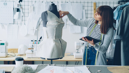 Young female fashion designing entrepreneur is working with tablet while checking clothing pinned...