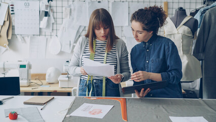 Creative clothing designers are looking at drawings and comparing them with images on tablet. Successful startup in fashion industry concept. Textiles, sewing machine and dummy are visible.