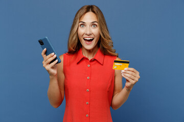 Young surprised shocked fun woman 30s wear red dress using mobile cell phone hold credit bank card...