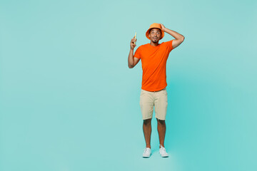 Full body young man of African American ethnicity wear orange t-shirt hat talk speak on mobile cell phone put hand on head isolated on plain pastel light blue cyan background People lifestyle concept