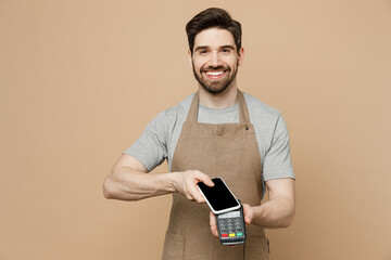 Young man barista barman employee in brown apron work in coffee shop hold mobile phone with blank screen bank payment terminal to pay isolated on plain beige background Small business startup concept.