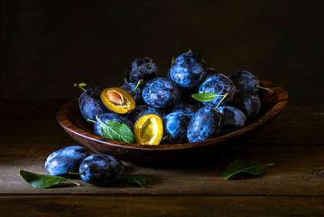 Ripe blue Fresh organic  sweet plums just picked from the tree, walnut wood bowl plate indoors. Autumn Harvest of fruits  rustic  wooden dark black background leaf clay.