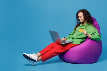Full body young woman of African American ethnicity in green shirt sit in bag chair use laptop pc computer credit bank card shopping online order delivery book tour isolated on plain blue background
