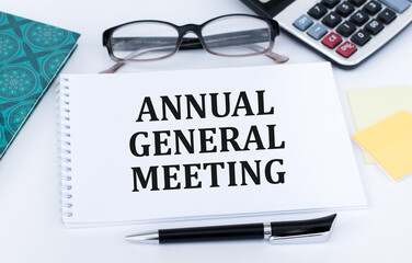 AGM Annual General Meeting is written in red on a white piece of paper on a light yellow background next to a laptop, pen, magnifying glass, glasses and a green plant.