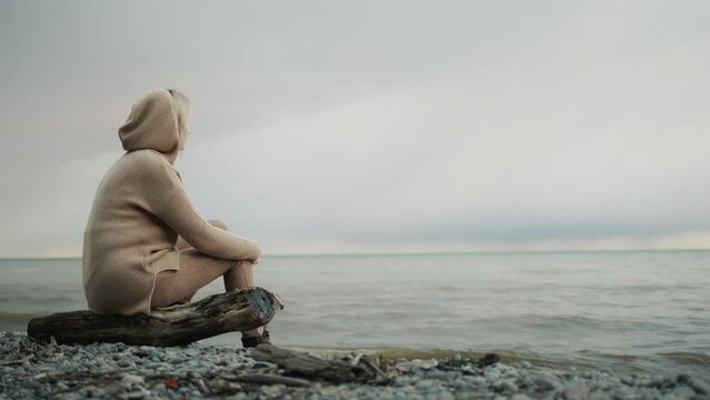 A woman in a warm sweater sits on a log on a rocky seashore where a storm begins