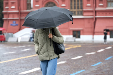 Rain in city, slim girl in jeans and jacket walk with black umbrella on a street. Rainy weather in...