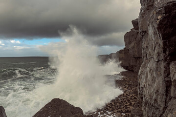 Ocean wave hits rock coastline of Ireland creating splash of water. Wild Irish nature scene. Burren, county Clare. Cloudy sky.