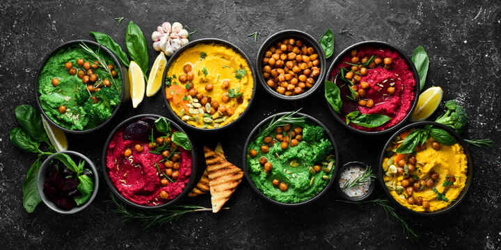 Colorful hummus bowls - green, yellow and beetroot hummus on dark background with lemon, olive oil, and spices. On a concrete black background.