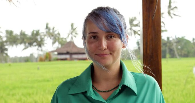 Pretty young woman with blue hair looks straight into the camera and smiles, we see that she wears a brackets on her teeth. Portrait shot with blurred background, green fields of Bali seen behind