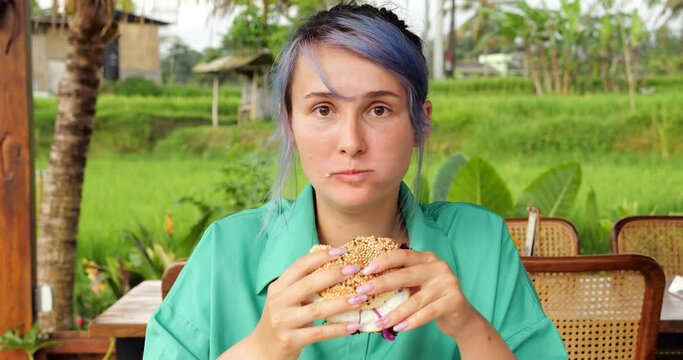 Pretty cheeky young woman with blue hair eating a big beef burger, looking straight at the camera. Green rice fields seen blurred on background, tourist girl enjoy high-calorie meal at Bali cafe