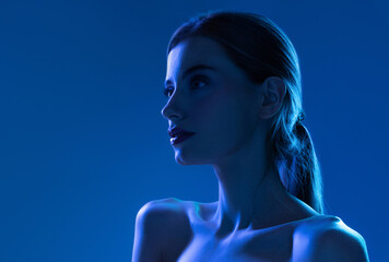Fototapeta premium Portrait of female fashion model in neon light on blue gradient studio background. Beautiful caucasian girl with trendy make-up and well-kept skin.