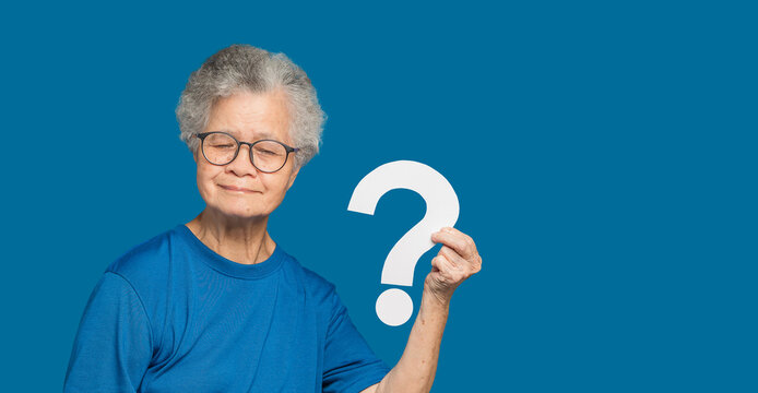 An elderly Asian woman holding a white question mark paper with a smile while standing on a blue background