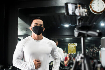 A muscular asian man wearing a white sweatshirt and a black facemask records a fitness vlog giving...