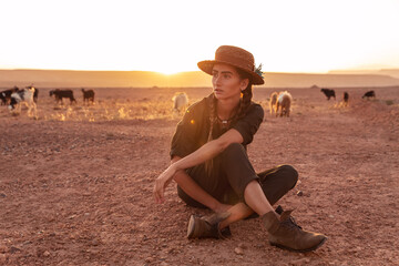 A girl in a hat sits on the sand in the desert at sunset. Goats grazing in the background. Morocco.