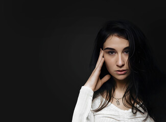 Portrait of young beautiful woman with long black hair and dark eyes, looking at camera and holds hand at temple of head, isolated on black background. Concept of emotions, hair, cosmetics. Copyspace.