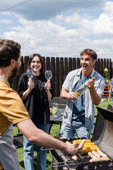 Cheerful man and woman holding wine and glasses near blurred friend cooking bbq in backyard