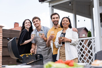 Smiling multiethnic couples holding glasses of wine and looking at camera near food on grill in backyard