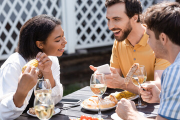 Smiling man holding smartphone and talking to interracial friends near food and wine during picnic