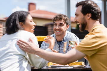 Smiling man looking at blurred multiethnic friends with wine near food during picnic outdoors