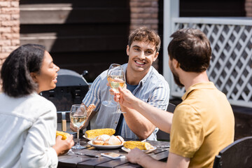 Fototapeta na wymiar Positive men clinking wine near food and blurred bi-racial friend outdoors