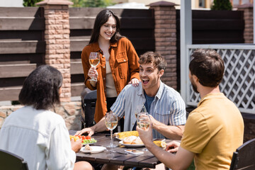 Positive interracial friends talking near tasty food and wine in backyard