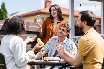 Positive woman holding glass of wine near blurred friends talking during picnic in backyard