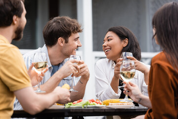 happy bi-racial woman holding glass of wine and looking at tattooed friend during bbq party