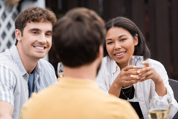 happy multiethnic couple with glasses of wine looking at friend on blurred foreground