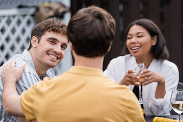 cheerful multiethnic friends smiling near blurred man on foreground during bbq party on backyard