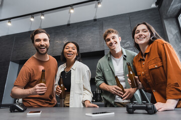 KYIV, UKRAINE - JULY 26, 2022: smiling multiethnic friends holding bottles of beer near joysticks and smartphones on kitchen worktop