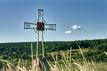 On a hill overgrown with wild grass there is a metal cross with the face of Jesus in the center, an Orthodox cross rises above the forest and valleys, a symbolic landscape