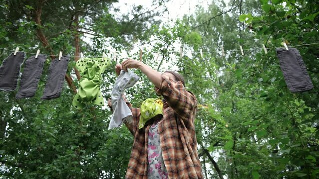 Middle age woman hanging reusable diapers to dry on clothesline in garden. Modern eco friendly cloth nappy for infant child hygiene. Sustainable lifestyle, zero waste concept.